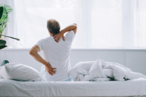 Man sitting on a bed holding his lower back and neck due to pain